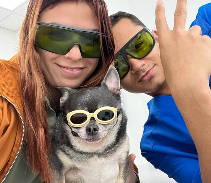 Dos personas y un pequeño chihuahua usando gafas de protección juntos en un interior.